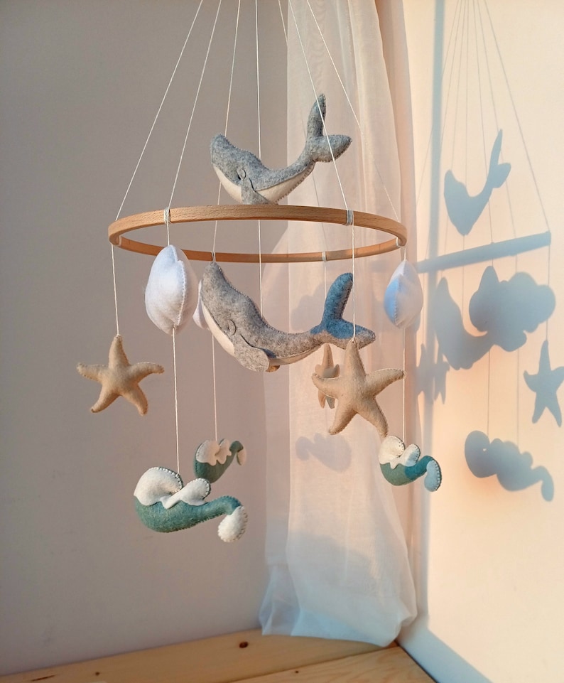 K&ouml;nnte beinhalten: Ein Holz-Babymobil mit maritimem Thema. Es zeigt zwei graue Wale, zwei Seesterne, zwei Wolken und zwei Wellen, alles aus Filz. Das Mobile h&auml;ngt an einer wei&szlig;en Decke.