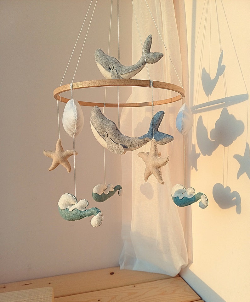 K&ouml;nnte beinhalten: Ein Holz-Babymobil mit maritimem Thema. Es zeigt zwei blaue und graue Wale, zwei beigefarbene Seesterne, zwei wei&szlig;e Wolken und zwei wei&szlig;e Eier. Das Mobile h&auml;ngt an einer wei&szlig;en Decke.
