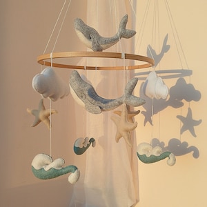 K&ouml;nnte beinhalten: Ein Holz-Babymobil mit maritimem Thema. Es zeigt zwei graue Filz-Wale, zwei wei&szlig;e Filz-Wolken, zwei blaue Filz-Wellen und zwei beige Filz-Seesterne.