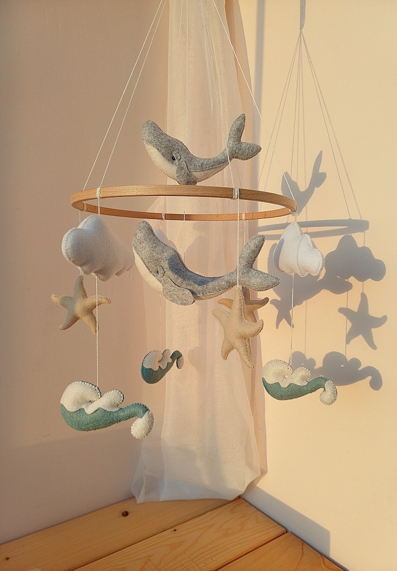 K&ouml;nnte beinhalten: Ein Holz-Babymobil mit maritimem Thema. Es zeigt zwei graue Filz-Wale, zwei wei&szlig;e Filz-Wolken, zwei blaue Filz-Wellen und zwei beige Filz-Seesterne.
