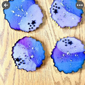 K&ouml;nnte beinhalten: Vier Untersetzer aus Kunstharz mit blau-violettem Aquarelldesign, jeweils mit goldenem Rand und kleinen schwarzen Meeresschildkr&ouml;ten-Silhouetten. Silberne Flocken sind &uuml;ber die Oberfl&auml;che verstreut. Die Untersetzer liegen auf einer Holzoberfl&auml;che.