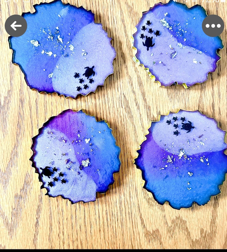 K&ouml;nnte beinhalten: Vier Untersetzer aus Kunstharz mit blau-violettem Aquarelldesign, jeweils mit goldenem Rand und kleinen schwarzen Meeresschildkr&ouml;ten-Silhouetten. Silberne Flocken sind &uuml;ber die Oberfl&auml;che verstreut. Die Untersetzer liegen auf einer Holzoberfl&auml;che.