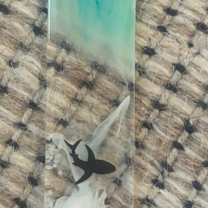 K&ouml;nnte beinhalten: Ein klarer Harz-Lesezeichen mit einer Strandszene. Die Oberseite hat ein t&uuml;rkis-wei&szlig;es Wirbeldesign. Ein schwarzer Hai, wei&szlig;e Objekte, ein Seestern, Muscheln und wei&szlig;er Sand sind in das Harz eingebettet.