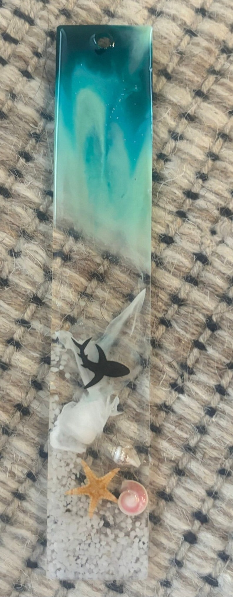 K&ouml;nnte beinhalten: Ein klarer Harz-Lesezeichen mit einer Strandszene. Die Oberseite hat ein t&uuml;rkis-wei&szlig;es Wirbeldesign. Ein schwarzer Hai, wei&szlig;e Objekte, ein Seestern, Muscheln und wei&szlig;er Sand sind in das Harz eingebettet.