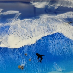 May include: Abstract ocean art piece with a shark silhouette. The artwork features a vibrant blue ocean with white foamy waves. Several small, textured rocks are scattered near the shark, adding depth to the scene. The overall effect is a dynamic and captivating depiction of the sea.