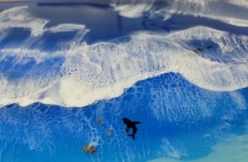 May include: Abstract ocean art piece with a shark silhouette. The artwork features a vibrant blue ocean with white foamy waves. Several small, textured rocks are scattered near the shark, adding depth to the scene. The overall effect is a dynamic and captivating depiction of the sea.