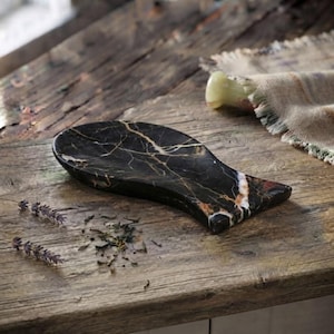 May include: A fish-shaped tray carved from black marble with prominent white and gold veining throughout the stone. The surface is smooth and polished, featuring a concave center and a distinct tail fin shape at one end of the object.