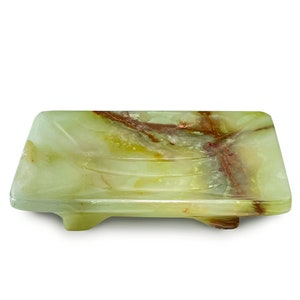 May include: A rectangular soap dish made of green onyx with brown and red veining. The dish has a slightly curved shape with three ridges and sits on four small feet. The natural stone has a polished finish.