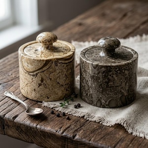 May include: Two cylindrical stone containers with matching lids, one in a light beige fossilized pattern and the other in a dark grey fossilized pattern, accompanied by a small silver-toned metal spoon placed on the side of the containers.