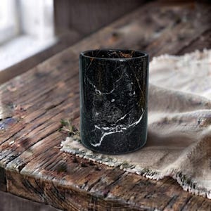 May include: A cylindrical container crafted from polished black marble, featuring prominent white and grey veining throughout its surface. The vessel has a smooth, vertical exterior and a circular opening at the top, showcasing the natural stone pattern.