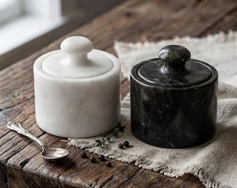 May include: Two cylindrical stone containers with matching lids, one in white marble and one in black marble, each featuring a rounded knob handle. A small silver-toned metal spoon rests nearby on a textured fabric surface.