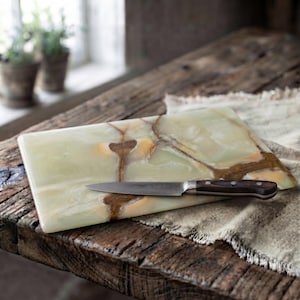 May include: A rectangular light green onyx stone serving board with natural brown veining patterns. A stainless steel paring knife with a dark brown wooden handle is placed horizontally across the center of the smooth, polished stone surface.