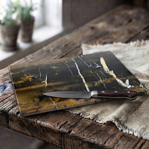 May include: A rectangular cutting board made of black and gold marble with prominent white veining. A kitchen knife with a dark brown wooden handle and a silver metal blade rests horizontally across the surface of the marble cutting board.