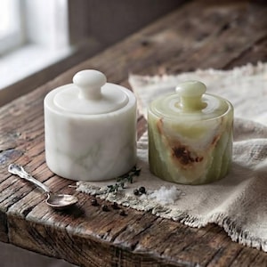 May include: Two cylindrical stone containers with matching lids and a small silver-toned metal spoon. One container is solid white with subtle grey veining, while the other is translucent green with brown mineral inclusions and a polished finish.