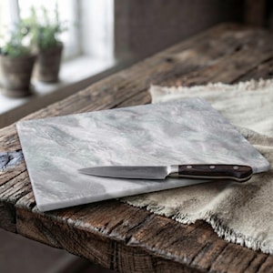 May include: A rectangular marble cutting board with a grey and white veined pattern. A kitchen knife with a dark brown wooden handle and a silver metal blade is positioned horizontally across the center of the stone surface.