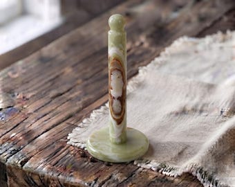 May include: A vertical paper towel holder crafted from polished green onyx stone. The stand features a circular base and a central cylindrical rod topped with a small sphere, showcasing natural banded patterns in shades of cream, brown, and light green.