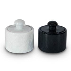 May include: Two small, round marble jars with lids. One jar is white with a textured surface, and the other is black and glossy. Both have a small, rounded knob on top of the lid. The jars are side by side on a white background.