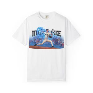 Pode incluir: Uma t-shirt branca com um gráfico de um lançador de basebol a lançar uma bola rodeada por efeitos de relâmpagos azuis. O jogador veste um uniforme branco com MIL 32 no peito. A palavra MILWAUKEE está impressa em letras pretas grandes acima.
