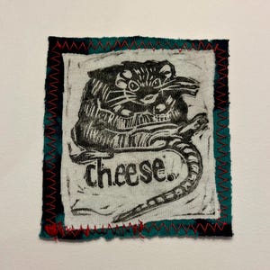 May include: A square fabric patch featuring a black and white illustration of a rat with the word "cheese" below. The patch is bordered by teal fabric with red stitching.