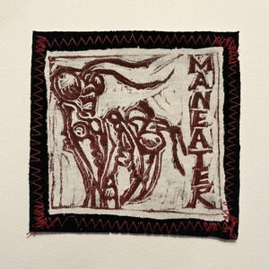 May include: Square fabric patch with a black border and red zigzag stitching. The central design features a maroon-colored abstract illustration and the word "MANEATER" in vertical block letters.