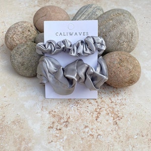 May include: Three silver satin scrunchies on a white card with the text "CALIWAVES". The card is on a background of light brown stones.