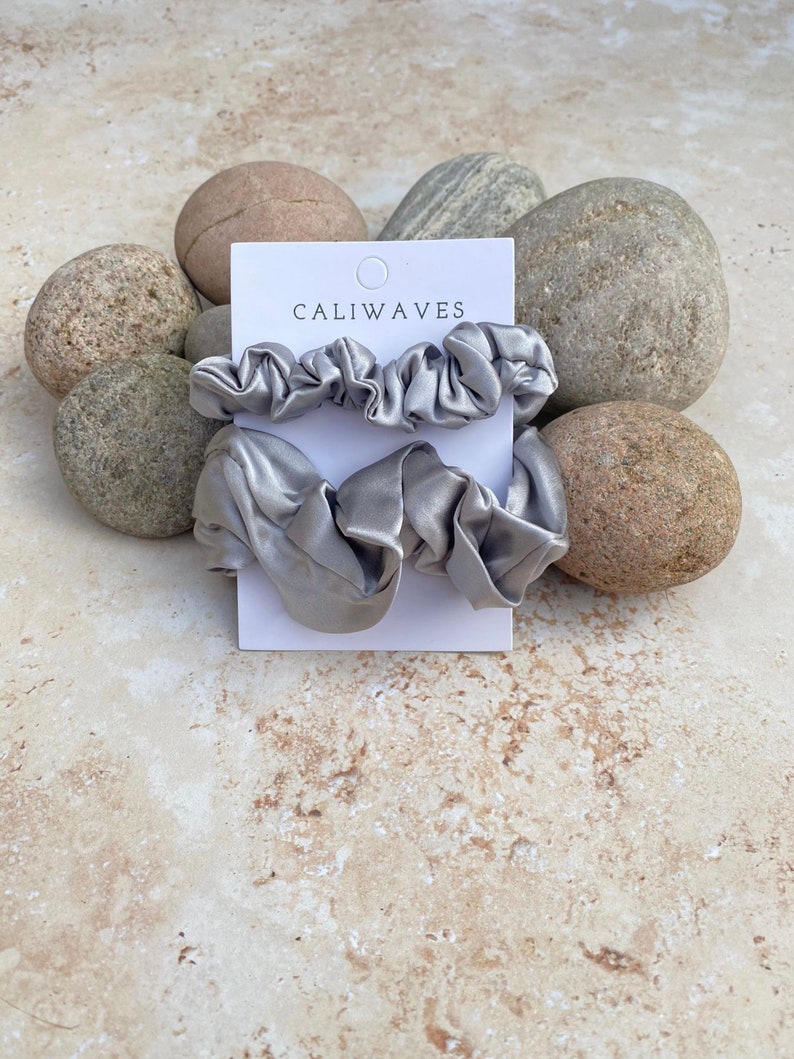 May include: Three silver satin scrunchies on a white card with the text "CALIWAVES". The card is on a background of light brown stones.