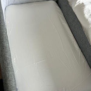 May include: A white mattress, protected by clear plastic, sits within a grey bed frame. The mattress is rectangular and appears unused. A white patterned headboard is visible in the background.