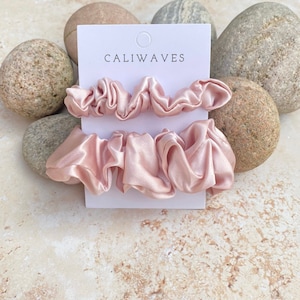 May include: Three pink satin scrunchies on a white card with the text "CALIWAVES" printed on it. The card is surrounded by rocks.