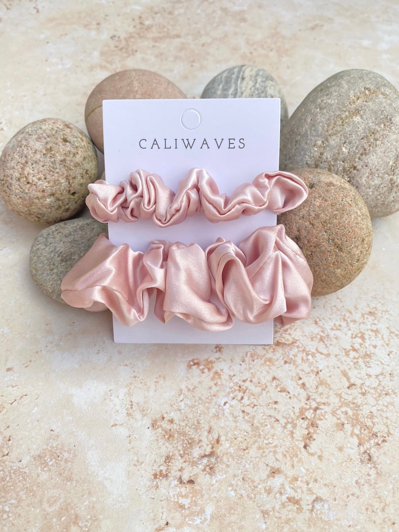 May include: Three pink satin scrunchies on a white card with the text "CALIWAVES" printed on it. The card is surrounded by rocks.