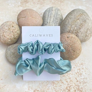 May include: Two light blue satin scrunchies on a white card with the text "CALIWAVES" printed on it. The card is sitting on a pile of light brown rocks.