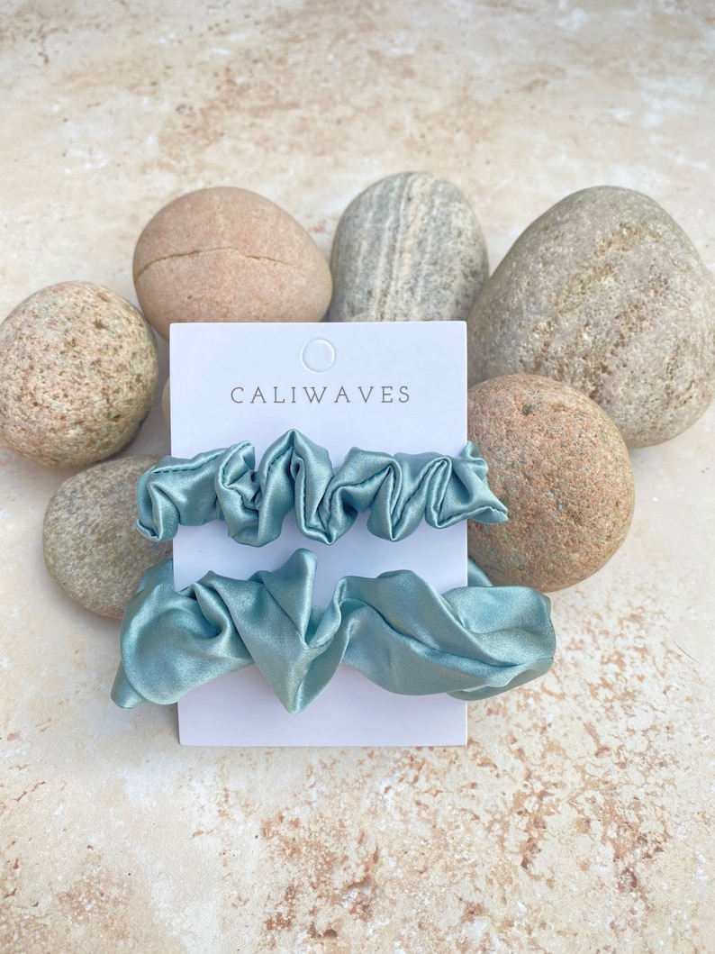 May include: Two light blue satin scrunchies on a white card with the text "CALIWAVES" printed on it. The card is sitting on a pile of light brown rocks.