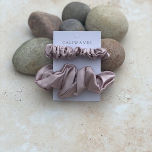 May include: Two dusty rose colored satin scrunchies on a white card with the text "CALIWAVES" printed on it. The card is sitting on a pile of gray and brown rocks.
