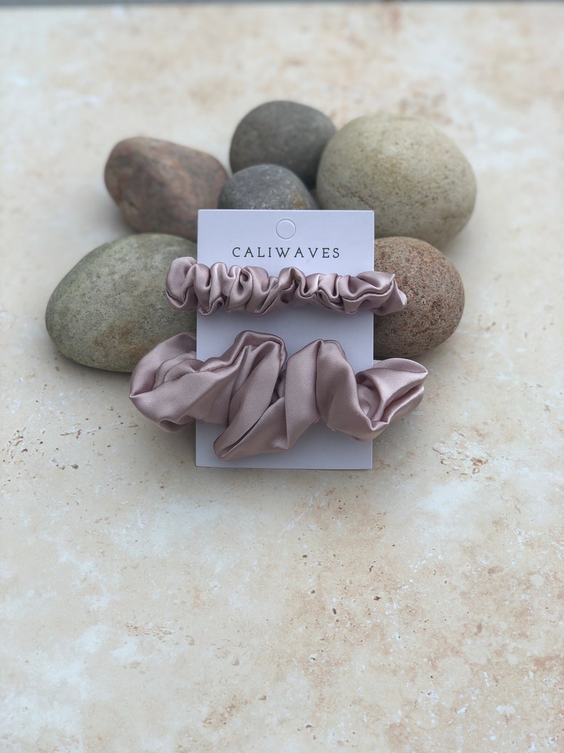 May include: Two dusty rose colored satin scrunchies on a white card with the text "CALIWAVES" printed on it. The card is sitting on a pile of gray and brown rocks.
