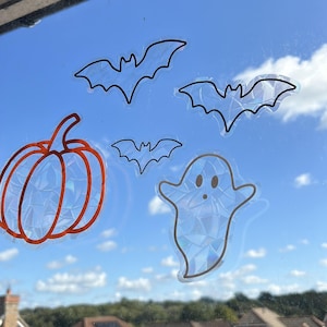 May include: Halloween window clings with a pumpkin, bats, and a ghost design. The pumpkin has an orange outline, and the bats and ghost have black outlines. The clings are iridescent, reflecting light, and are set against a blue sky.