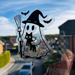 May include: A Halloween window sticker depicting a ghost in a witch's hat, holding a broom, with a black cat and bats. The cling has an iridescent, textured finish. The design is outlined in black, creating a decorative effect for the window.