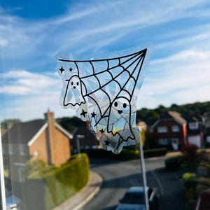 May include: A Halloween window cling with a spider web design, two smiling ghost figures, and star accents. The cling has a transparent, iridescent finish with black outlines. The background shows a blue sky and houses in a residential area.
