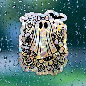 May include: A holographic ghost sticker featuring a bow, bat, spiderweb, flowers, and jack-o'-lanterns. The ghost has large black eyes and is surrounded by a floral and Halloween-themed design. The sticker has a mosaic-like appearance.