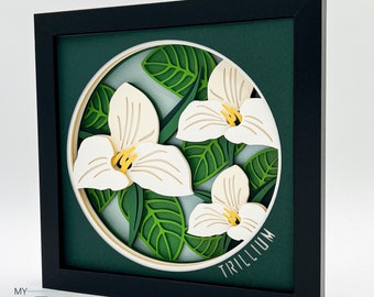 Puede incluir: Una obra de arte de papel enmarcada en negro con un fondo blanco y un círculo en el centro. El círculo presenta un diseño de corte de papel 3D de flores de trillium blancas con hojas verdes. El texto "TRILLIUM" está escrito en negro en la parte inferior del círculo.