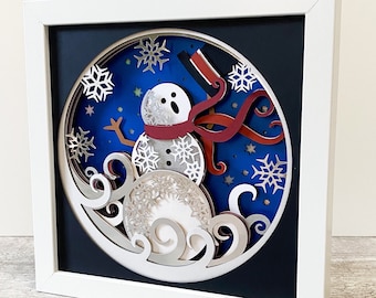 May include: A white frame with a blue background and a 3D paper cut-out of a snowman wearing a red scarf and a black hat. The snowman is surrounded by white snowflakes and swirls.