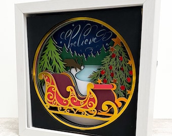 May include: A white frame with a black background and a gold circle containing a 3D paper cut-out design of a sleigh with a red and gold design, a Christmas tree with red ornaments, and a forest scene. The text "Believe" is written in cursive on the circle.