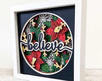 May include: A layered 3D Christmas-themed artwork in a white frame. The artwork features a circular design with red poinsettias, green foliage, and the word "believe" in white script. Snowflakes and berries add to the festive look.