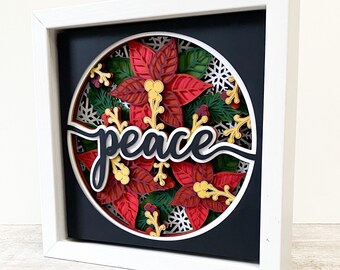 May include: A white frame with a black background and a round cutout featuring a 3D papercraft design of red poinsettia flowers, green leaves, and white snowflakes. The word "peace" is written in black cursive script.