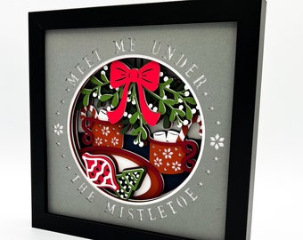 May include: A 3D papercraft scene with a black frame. The scene depicts a festive holiday setting with a red bow, green leaves, and two mugs with marshmallows. The text "Meet me under the mistletoe" is written in a circle around the scene.
