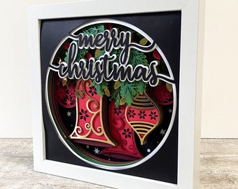 May include: A white frame with a black background and a circular cutout featuring a 3D papercraft design of a red and green Christmas ornament and bells with the text "merry christmas" in black.