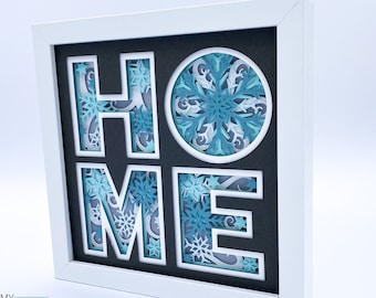 May include: A white frame with a black background and a 3D paper cut design of the word "HOME" with a snowflake pattern. The letters are layered and have a shadow effect.