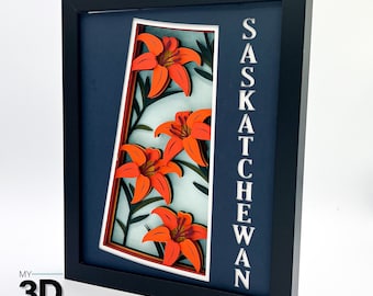 May include: A framed paper art piece featuring a stylized outline of the province of Saskatchewan, Canada, with the word "Saskatchewan" printed vertically on the right side. The outline is filled with a layered paper design of orange lilies.