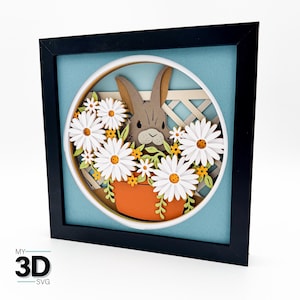 May include: A framed 3D paper art piece featuring a brown bunny surrounded by white daisies with orange centers and small yellow flowers. The bunny is in an orange pot, set against a light blue background, with a white lattice fence. The frame is black.