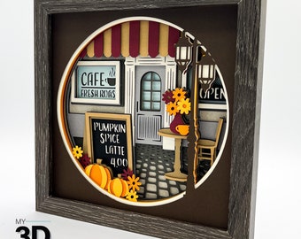 May include: A 3D papercraft scene of a cafe with a red and white awning, a sign that says "Cafe Fresh Roasts", and a chalkboard sign that says "Pumpkin Spice Latte 4.00". The scene is set in a fall theme with pumpkins, flowers, and a street lamp.