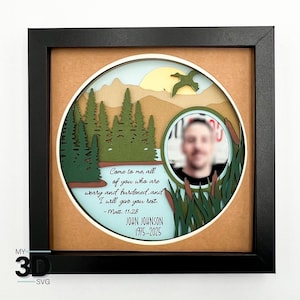 Op de afbeelding: Een ingelijst 3D-papierkunstwerk met een zwart frame. Het kunstwerk toont een gelaagd landschap met een meer, bomen, bergen en een zon. Een cirkelvormige foto is inbegrepen, samen met de tekst: "Komt tot Mij, allen die vermoeid en belast zijt, en Ik zal u rust geven."