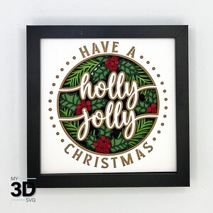 May include: A black framed picture with a white background and a gold circle with the text "Have a holly jolly Christmas" in white lettering. The circle is surrounded by green holly leaves and red berries.